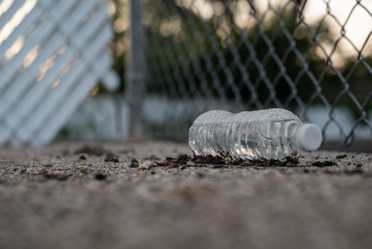 Close-up of a plastic water bottle discarded on gravel near a chain-link fence.